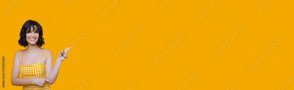 Lovely caucasian woman wearing a dress is pointing to something with her index finger and smiling cheerfully on a yellow blank space Lovely caucasian woman wearing a dress is pointing to something with her index finger and smiling cheerfully on a yellow blank space