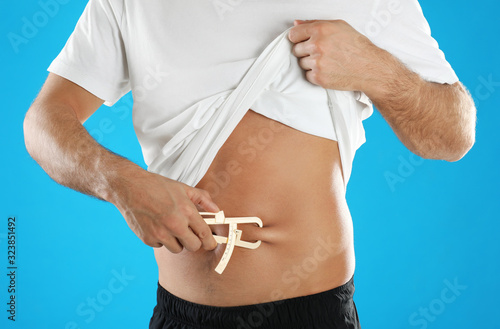 Man measuring body fat layer with caliper on light blue background, closeup. Nutritionist's tool