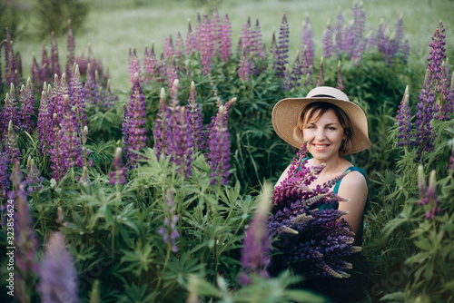 A beautiful romantic woman smiles joyfully in a blue dress and hat sits in a flower field with a huge bouquet of purple lupine flowers. Soft selective focus.