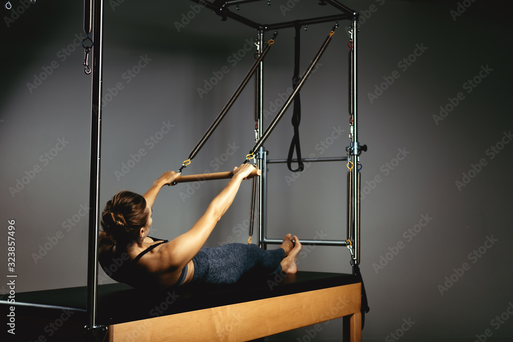 Fototapeta A young girl does Pilates exercises with a bed reformer, barrel machine tool. Beautiful slim fitness trainer on the background of a reformer doing various exercises gray, low key, light art. Fitness