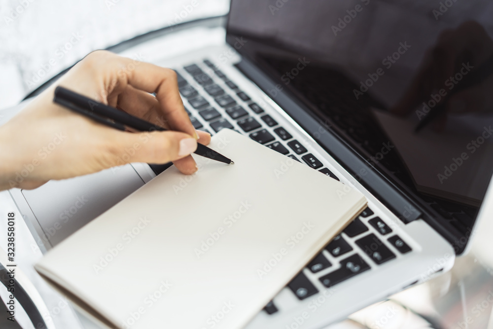 Woman writes with a pen in notebook on laptop keyboard in a sunny office, business and education concept. Close up Woman writes with a pen in notebook on laptop keyboard in a sunny office, business and education concept. Close up