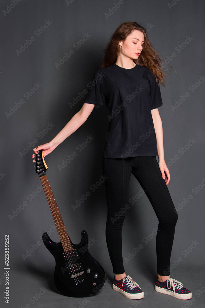 Attractive, young rock girl in black clothes with an electric guitar in her hands stands on a gray background. Model with clean skin. Attractive, young rock girl in black clothes with an electric guitar in her hands stands on a gray background. Model with clean skin.