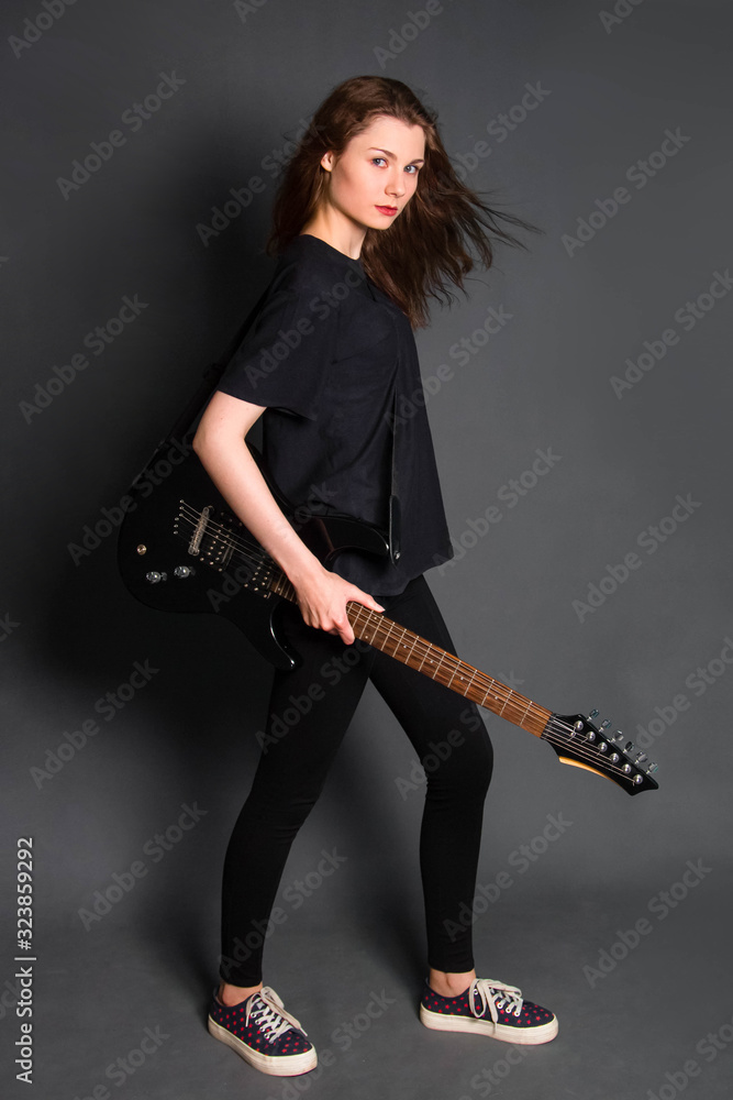 Attractive, young rock girl in black clothes with an electric guitar in her hands stands on a gray background. Model with clean skin. Attractive, young rock girl in black clothes with an electric guitar in her hands stands on a gray background. Model with clean skin.