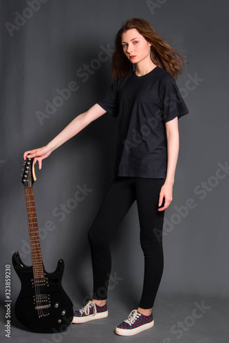 Attractive, young rock girl in black clothes with an electric guitar in her hands stands on a gray background. Model with clean skin.