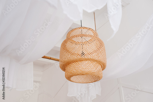 A stylish wicker wooden chandelier in boho and Bali style hangs on the ceiling near a white canopy made of weightless tulle. Part of the light airy interior.