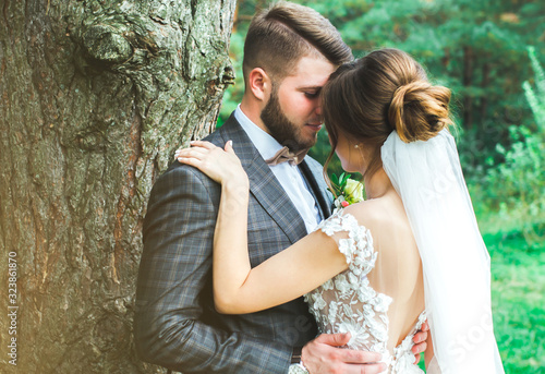 Beautiful wedding couple in the forest. The bride with tulle veil and open low back elegant dress is hugging the groom in bow tie. Wedding buttonhole and checkered suit in Great Gatsby style.