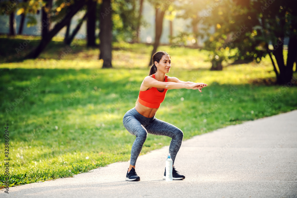 Full length of positive fit sportswoman doing squats endurance while standing on path in nature. Full length of positive fit sportswoman doing squats endurance while standing on path in nature.