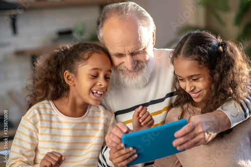 Grey-haired bearded man and his granddaughters watching a video together and smiling