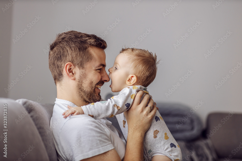 Adorable little blond boy playing with his caring father and trying to bite his nose. Family values. - obrazy, fototapety, plakaty 