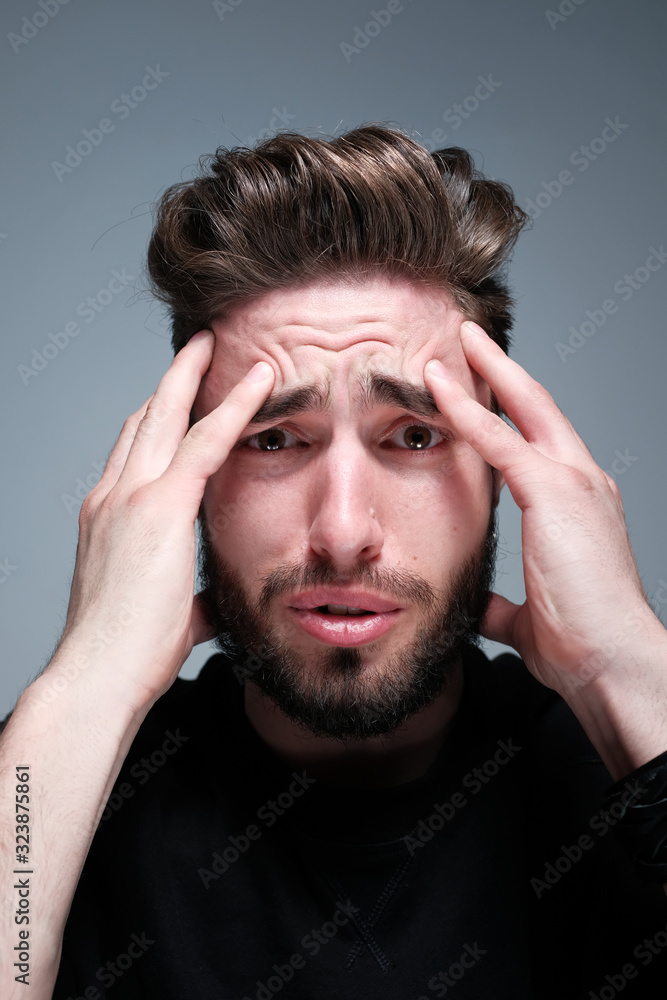 A young dark-haired man with a beard holds his head and shows different human emotions: hatred, fear, despair, horror, malaise, headache, clairvoyance. Close-up studio portrait of a man. A young dark-haired man with a beard holds his head and shows different human emotions: hatred, fear, despair, horror, malaise, headache, clairvoyance. Close-up studio portrait of a man.