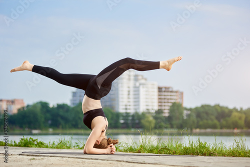 Young woman practicing Salamba Sirsasana exercise, supported headstand pose at the city lake in the morning, leading healthy lifestyle, summer vibes