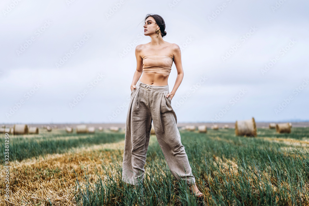 Brunette in fashionable clothes with bare shoulders standing in a wheat field on green grass. Woman holds her hands in pants pockets and looks away Brunette in fashionable clothes with bare shoulders standing in a wheat field on green grass. Woman holds her hands in pants pockets and looks away