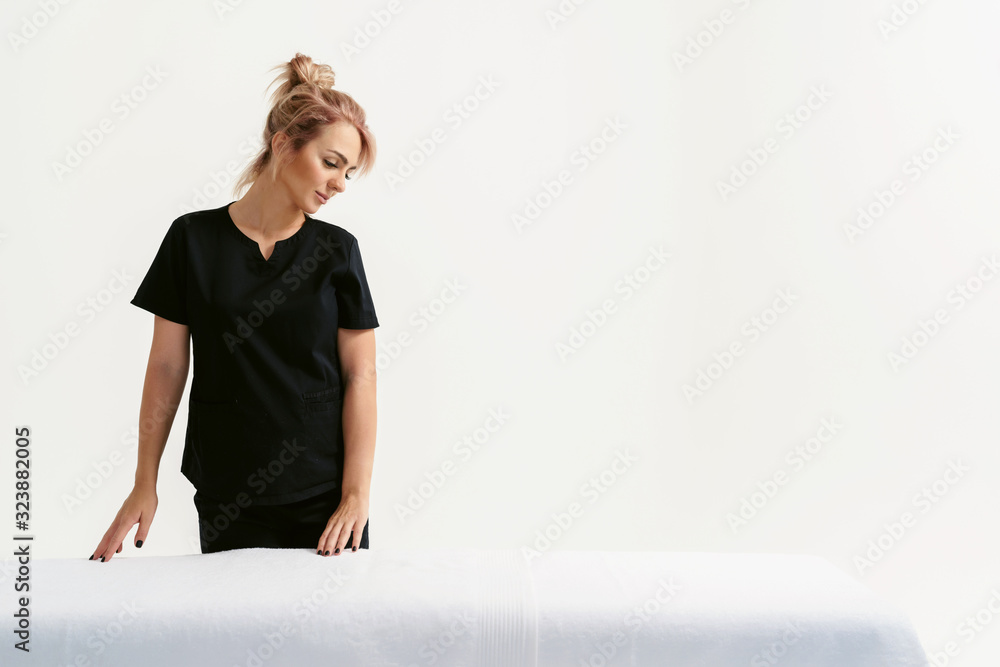 Portrait of young smiling friendly funny woman masseur in uniforme near massage table in Spa Salon. Physical Therapist In Medical Office. Beauty Treatment, Massage Therapy. White background isolated - obrazy, fototapety, plakaty 