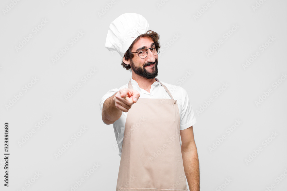 young crazy chef pointing at camera with a satisfied, confident, friendly smile, choosing you against white wall - obrazy, fototapety, plakaty 