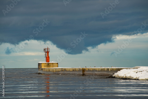 The Baltic sea at cold winter cloudy day. The gate to the port. Seagulls on the lighthouse and on the breakwater. Snowy pier. Liepaja, Latvia.