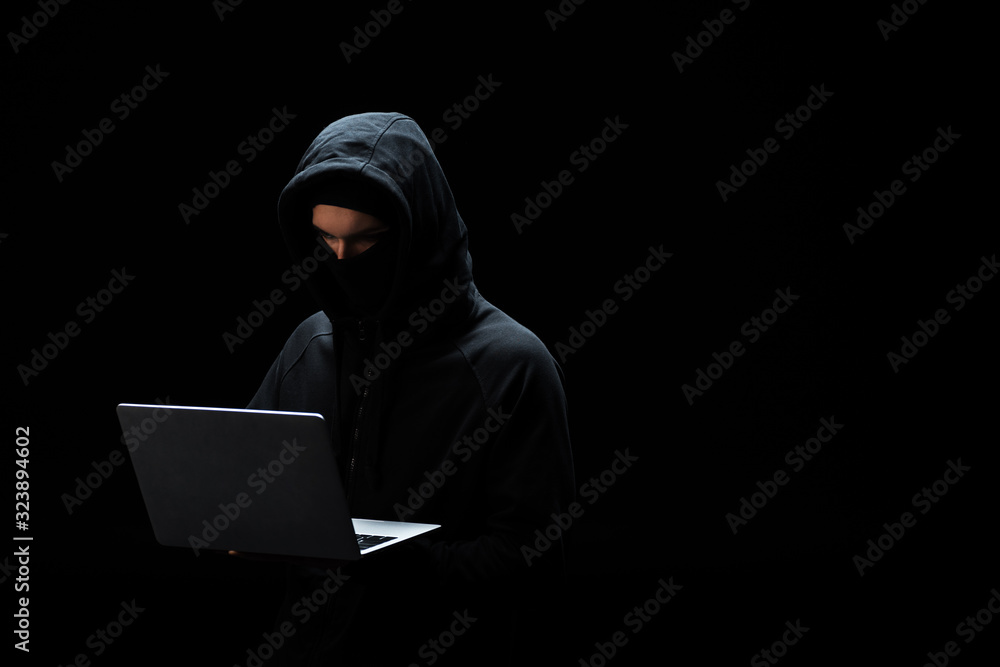 hacker in mask and hood using laptop isolated on black