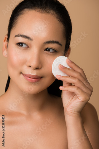 smiling beautiful naked asian girl holding cotton pad on face isolated on beige
