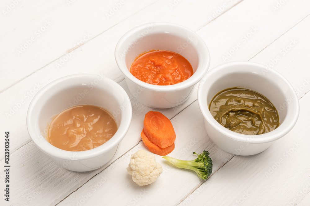 Bowls with baby nutrition with carrot slices and pieces of cauliflower and broccoli on white wooden background Bowls with baby nutrition with carrot slices and pieces of cauliflower and broccoli on white wooden background