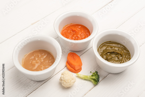 Bowls with baby nutrition with carrot slices and pieces of cauliflower and broccoli on white wooden background