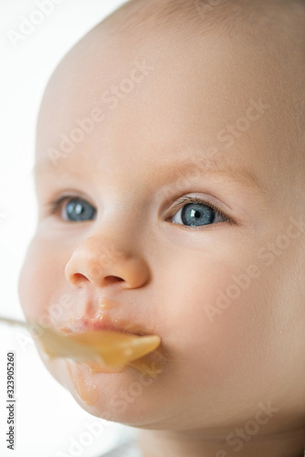 Selective focus of cute baby looking away during feeding with fruit puree isolated on white