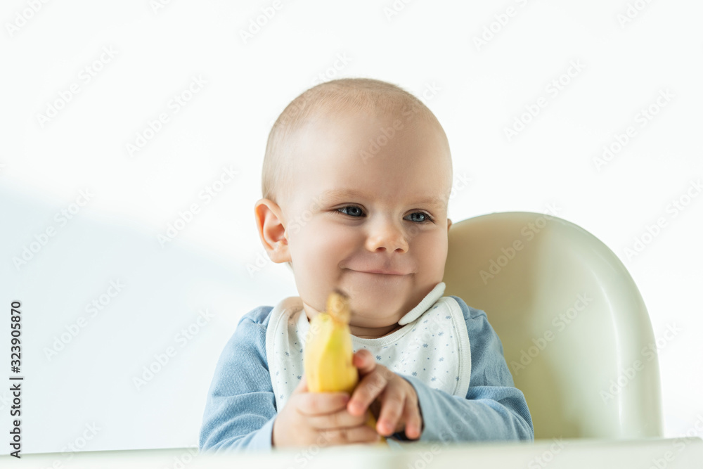 Selective focus of smiling baby holding banana while sitting on feeding chair on white background Selective focus of smiling baby holding banana while sitting on feeding chair on white background