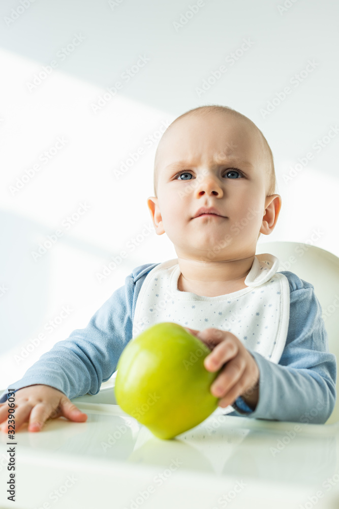 Selective focus of pensive baby boy holding green apple while sitting on feeding chair on white background Selective focus of pensive baby boy holding green apple while sitting on feeding chair on white background