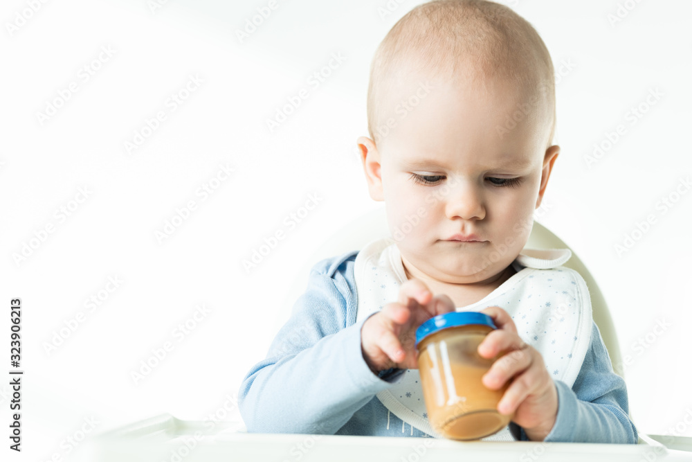 Selective focus of baby boy on feeding chair holding jar with fruit puree on white background