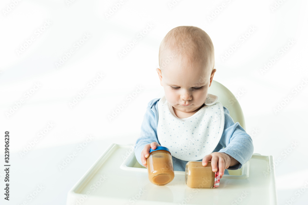 Adorable baby boy holding jars with baby food on table of feeding chair on white background Adorable baby boy holding jars with baby food on table of feeding chair on white background