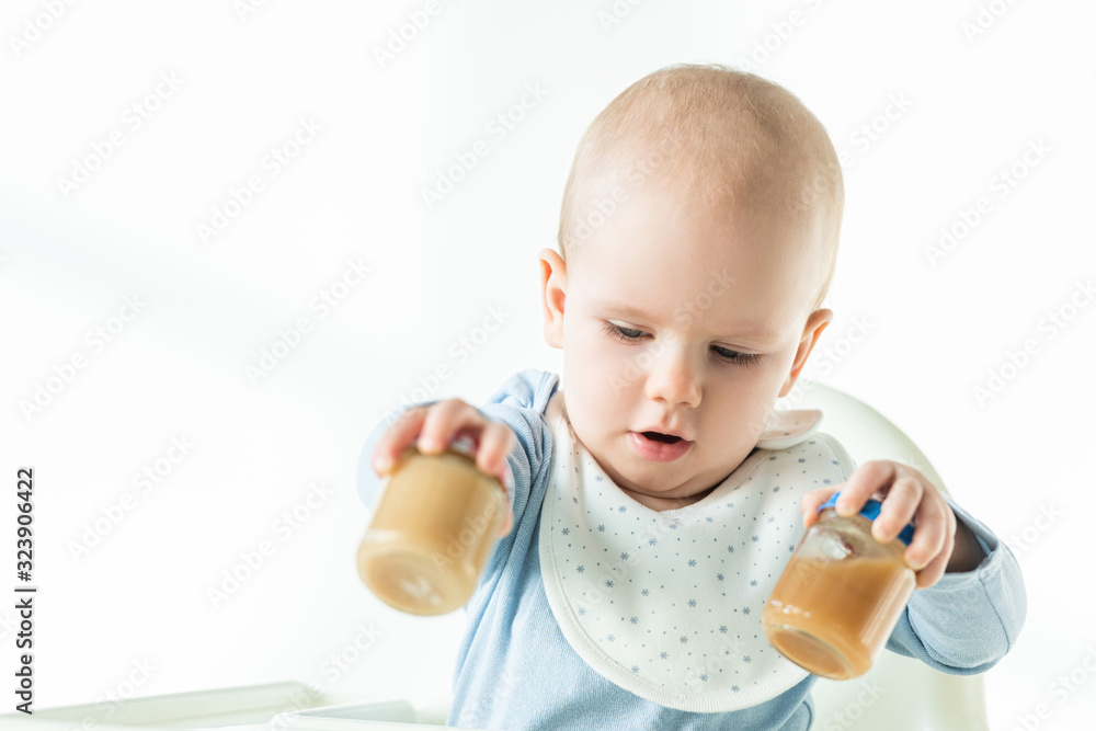 Selective focus of infant on feeding chair holding jars of fruit baby nutrition on white background Selective focus of infant on feeding chair holding jars of fruit baby nutrition on white background