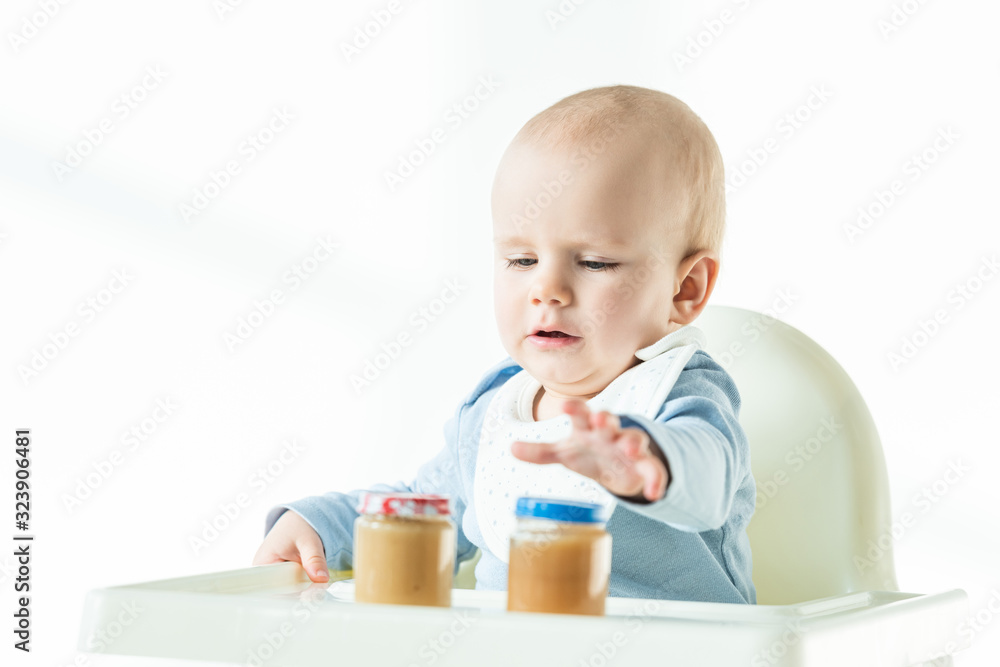 Selective focus of baby sitting on feeding chair with jars on fruit puree on table on white background Selective focus of baby sitting on feeding chair with jars on fruit puree on table on white background
