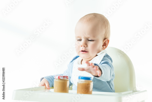 Selective focus of baby sitting on feeding chair with jars on fruit puree on table on white background