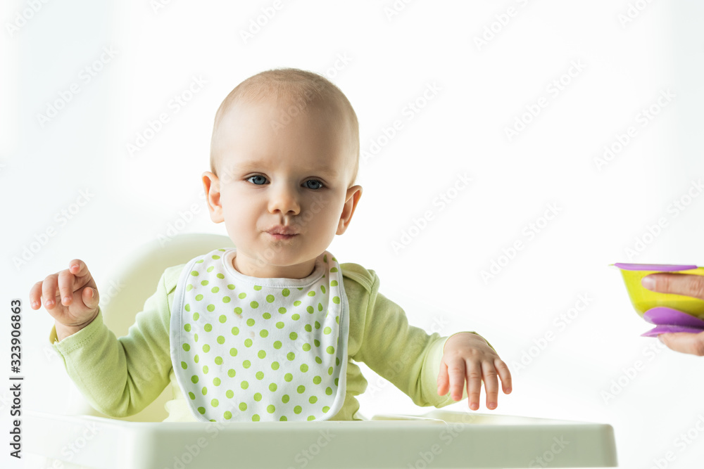 Adorable baby looking at camera while sitting on feeding chair near mother with bowl of baby nutrition on white background Adorable baby looking at camera while sitting on feeding chair near mother with bowl of baby nutrition on white background