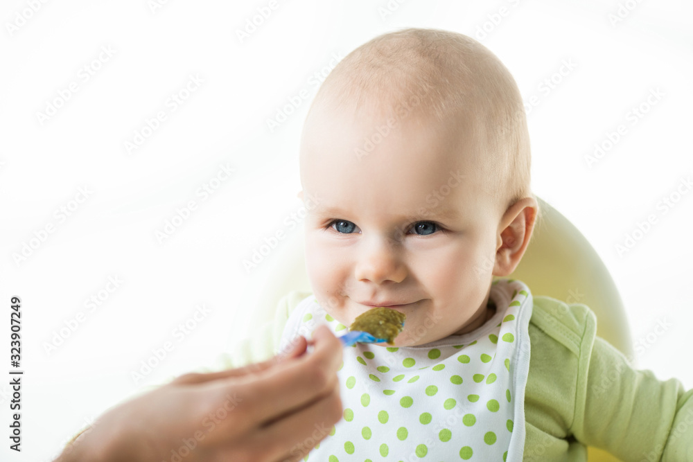 Selective focus of infant looking at mother with spoon while eating on feeding chair isolated on white Selective focus of infant looking at mother with spoon while eating on feeding chair isolated on white