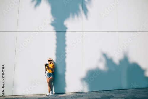 Trendy woman in casual wear using photo camera while leaning on wall at city street