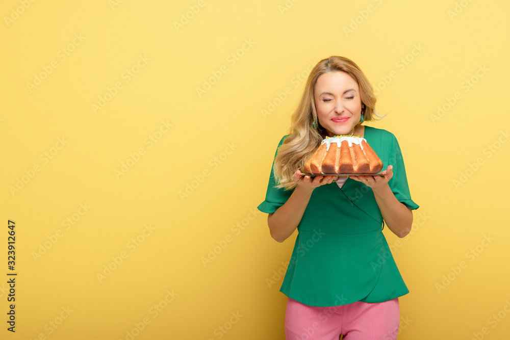 smiling woman with closed eyes holding tasty easter cake isolated on yellow