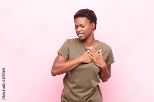 young pretty black womanlooking sad, hurt and heartbroken, holding both hands close to heart, crying and feeling depressed against pink wall photo