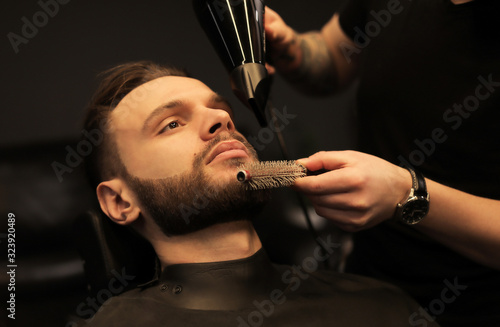 Beard styling. Low angled shot of barber’s hands holding hairdryer and small round comb while styling beard of his customer.