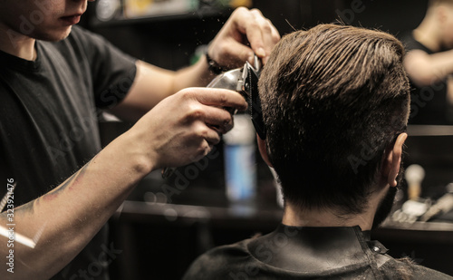 Creative process. Side view photo of a young barber trimming his customer’s hair with an electric shaver and a comb.