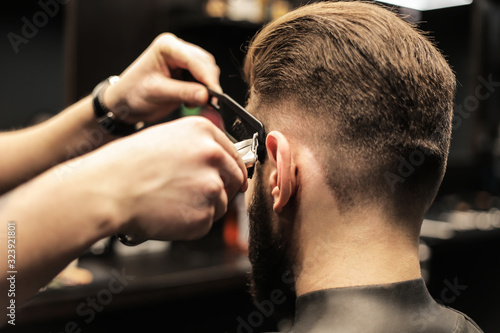 Making a new haircut. Close up cropped shot of barber’s hands trimming hair of his customer with electric razor and a special comb.