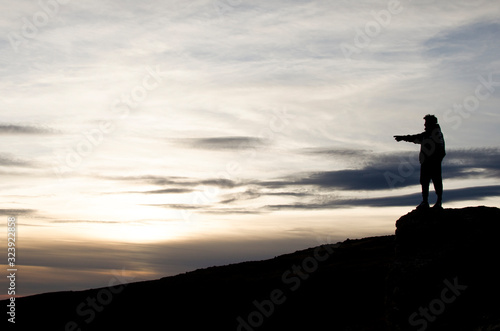 man on top of a mountain points the horizon towards a mountain landscape at sunset