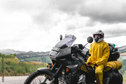 Fototapeta Naklejka Na Ścianę i Meble -  Motorcyclist traveler girl wearing yellow raincoat and sitting in a motorcycle saddle. Motorbike on mountain road. Extreme travel tour. Biker equipment. Dramatic cloud, copy space