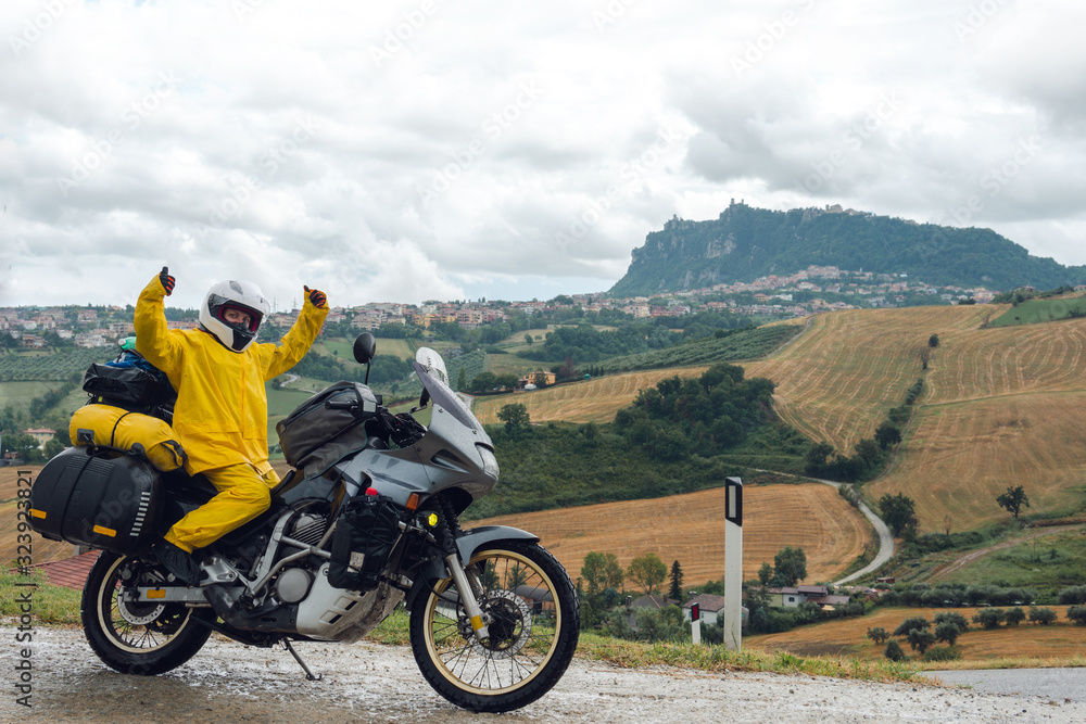Happy motorcyclist traveler girl wearing yellow raincoat and sitting in a motorcycle saddle. Destination scene. Extreme travel tour. Biker equipment. View of the mountain. San Marino, Italy. Happy motorcyclist traveler girl wearing yellow raincoat and sitting in a motorcycle saddle. Destination scene. Extreme travel tour. Biker equipment. View of the mountain. San Marino, Italy.