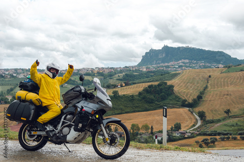 Happy motorcyclist traveler girl wearing yellow raincoat and sitting in a motorcycle saddle. Destination scene. Extreme travel tour. Biker equipment. View of the mountain. San Marino, Italy.