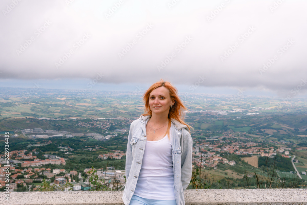 Portrait of a blonde girl. Dressed in a light denim jacket, blue pants and a white tank top. Posing on top of a mountain overlooking the city of San Marino, Italy