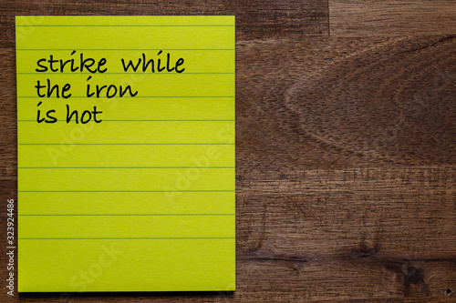 yellow sticker with lines on a dark wooden background with the handwriting of the wisdom of life: strike while the iron is hot