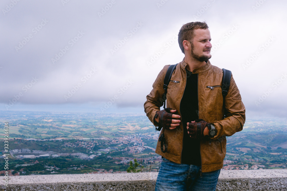 Portrait of a man dressed in a stylish brown leather jacket and jeans. backpack watches and biker gloves. posing on top of a mountain with a view of the city. Copy space Portrait of a man dressed in a stylish brown leather jacket and jeans. backpack watches and biker gloves. posing on top of a mountain with a view of the city. Copy space