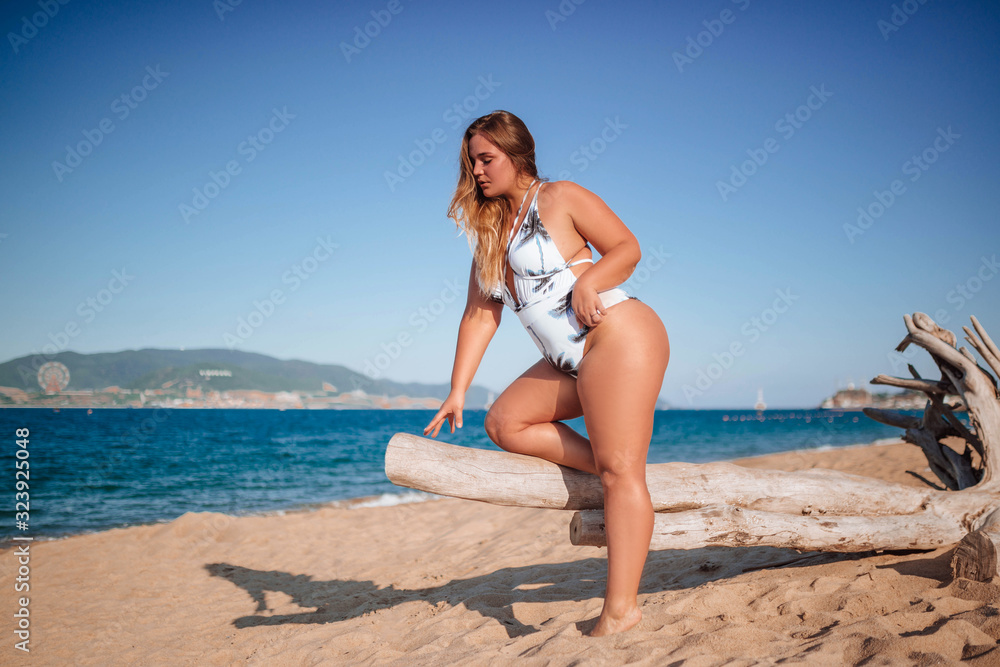 Girl, xxl size model, plump girl in a white one-piece swimsuit on a sandy beach posing near palm trees
