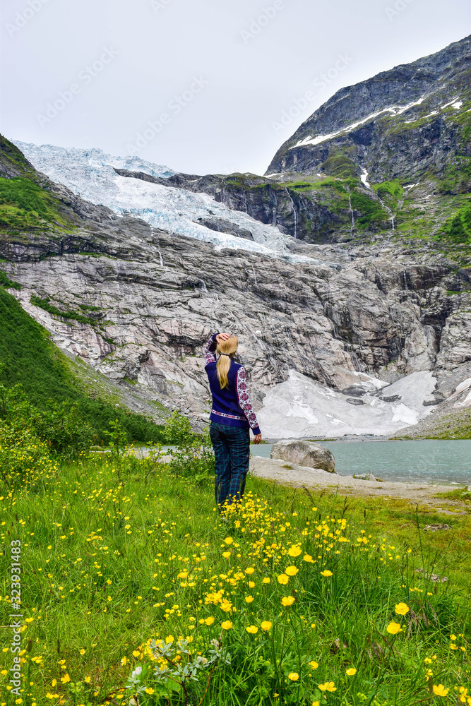 The young girl looks at the Boyabreen glacier, which is the sleeve of large Jostedalsbreen glacier and wild yellow flowers in the foreground. Melting glacier forms the lake with clear water. Norway. The young girl looks at the Boyabreen glacier, which is the sleeve of large Jostedalsbreen glacier and wild yellow flowers in the foreground. Melting glacier forms the lake with clear water. Norway.