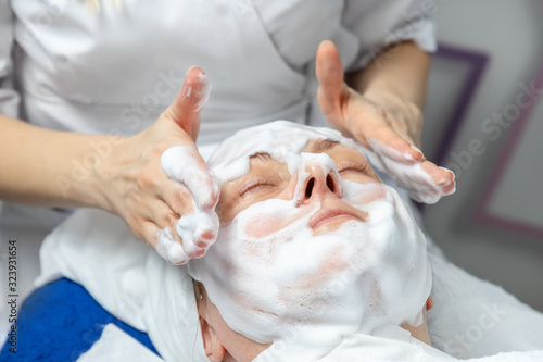 Close-up beautician doctor hands making anti-age procedures  applying foam cleansing mask for mid-aged female client at beauty clinic. Cosmetologist doing skincare treatment .Health care therapy