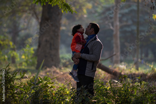 An Indian brunette father and his baby boy in winter garments enjoying themselves in winter afternoon in light and shadow in green forest background. Indian lifestyle and parenthood.